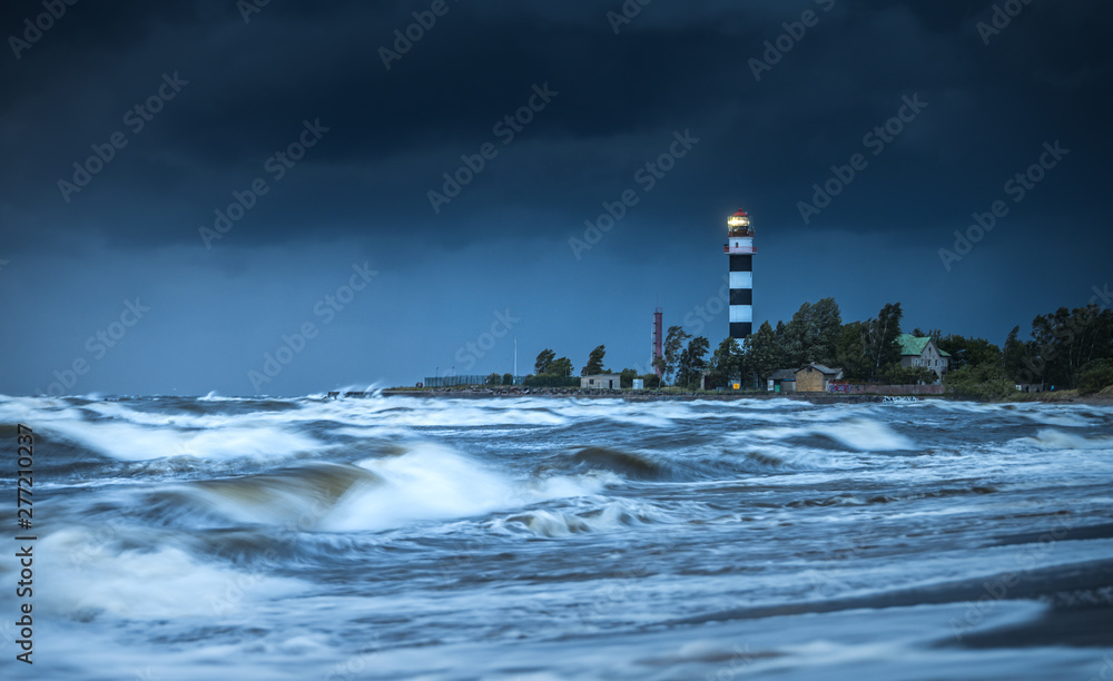 Thunderstorm over Baltic sea in a windy summer evening. Impressive waves hitting breakwater. Lighthouse covered in dark storm clouds above. Sandy seashore covered in tall grass.