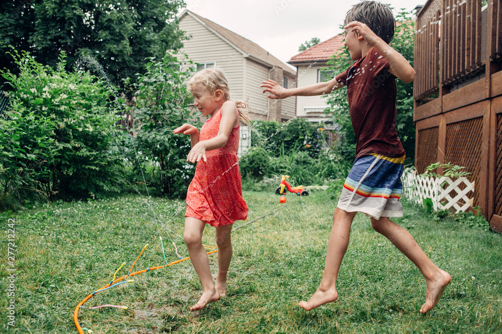 Fototapeta premium Kids friends boy and girl splashing with gardening hose sprinkle on backyard on summer day. Children playing with water outside at home yard. Candid authentic real life moment of funny family activity