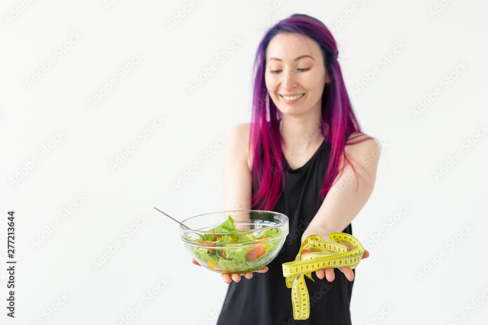 Young asian girl hipster colored hair holding in hands a measuring tape and vegetable salad posing on a white background with copyspace. Healthy eating concept. Young asian girl hipster colored hair holding in hands a measuring tape and vegetable salad posing on a white background with copyspace. Healthy eating concept.