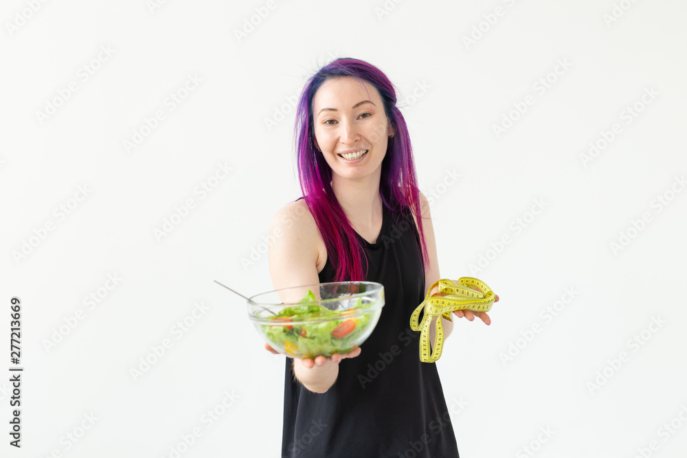 Young asian girl hipster colored hair holding in hands a measuring tape and vegetable salad posing on a white background with copyspace. Healthy eating concept. Young asian girl hipster colored hair holding in hands a measuring tape and vegetable salad posing on a white background with copyspace. Healthy eating concept.