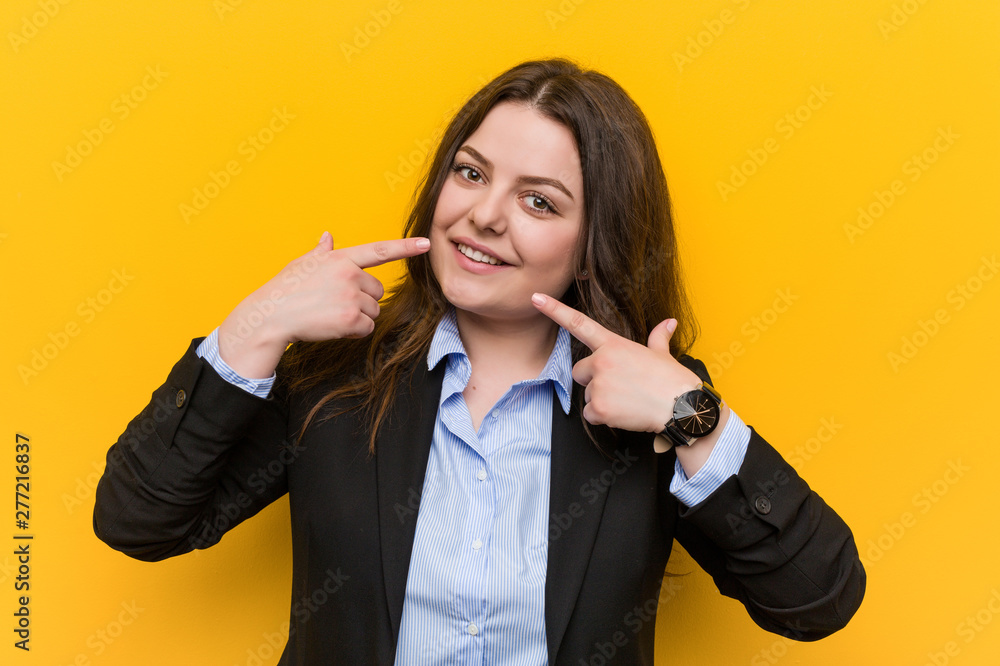 Young plus size caucasian business woman smiles, pointing fingers at mouth.
