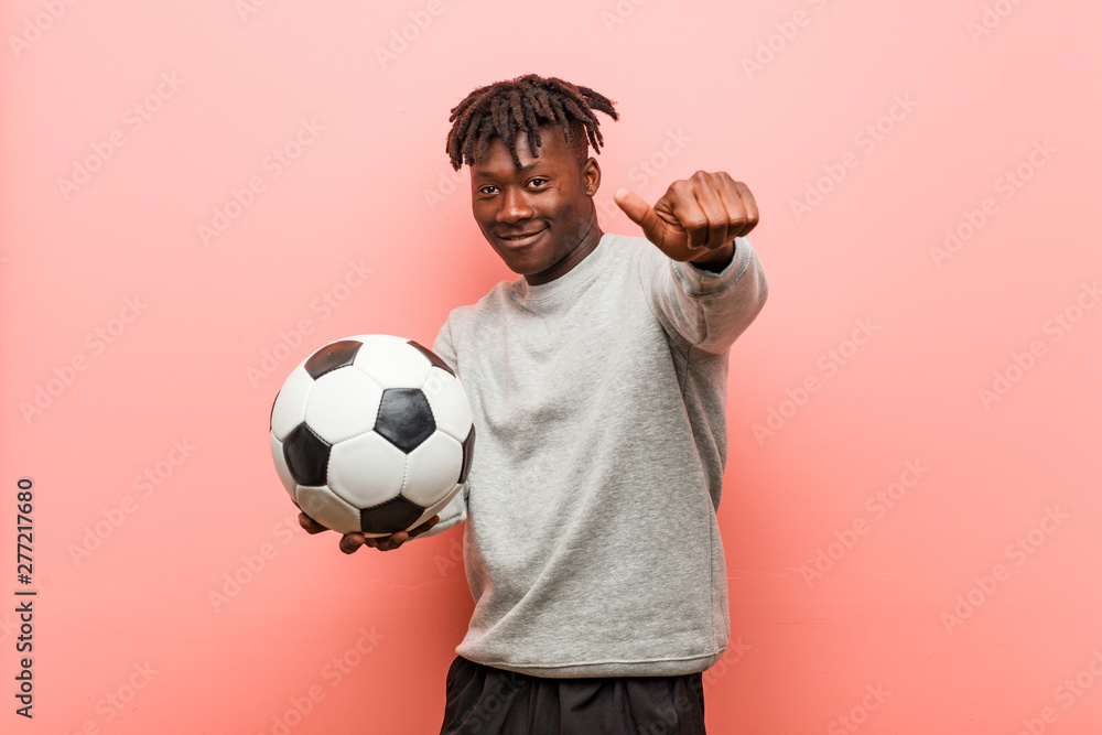 Young fitness black man holding a soccer ball cheerful smiles pointing to front.