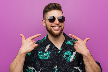 Young man wearing a vacation look smiles, pointing fingers at mouth.