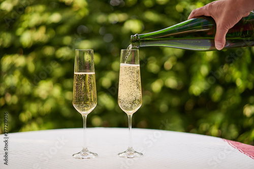 Pouring a glass of white sparkling wine or champagne into flute style wine glass with lot of bubbles, in the outdoor garden restaurant on the table with white tablecolth in sunny summer evening.