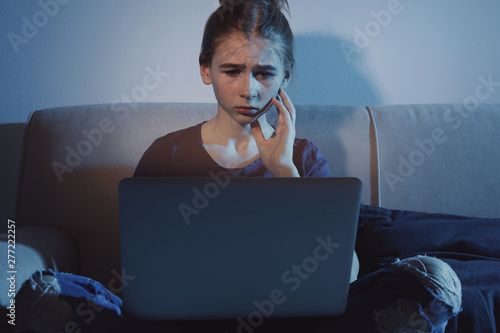 Upset teenage girl with laptop on sofa in dark room. Danger of internet photo