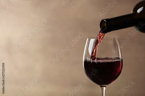 Pouring red wine into glass from bottle against blurred beige background, closeup. Space for text