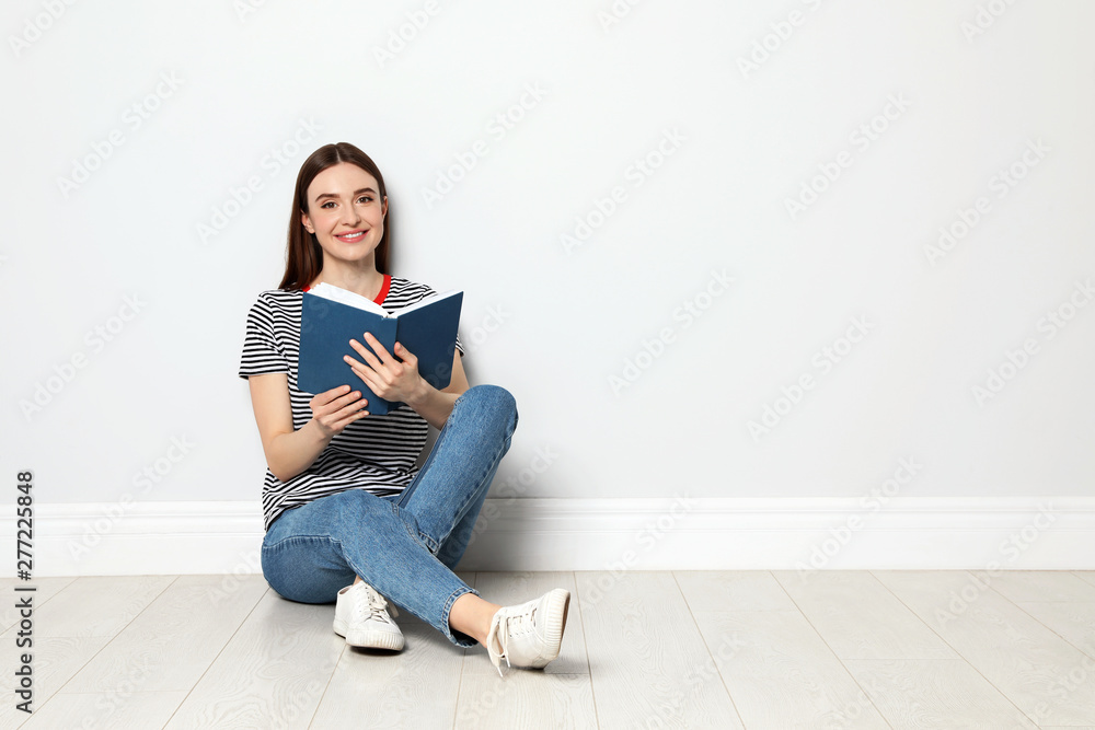 Young woman reading book on floor near white wall, space for text Young woman reading book on floor near white wall, space for text