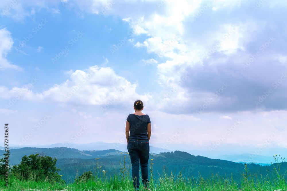 Woman with backpack looking at amazing woods and mountains in summer, travel concept, space for text. View of woods and blue sky and clouds on vacation