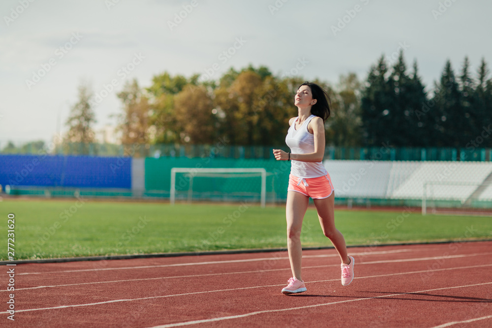 Sport. Athletic young brunette woman in pink sneakers, shorts and tops run on running track stadium at sunset. her hair is developing. Concept run. concept of a healthy lifestyle