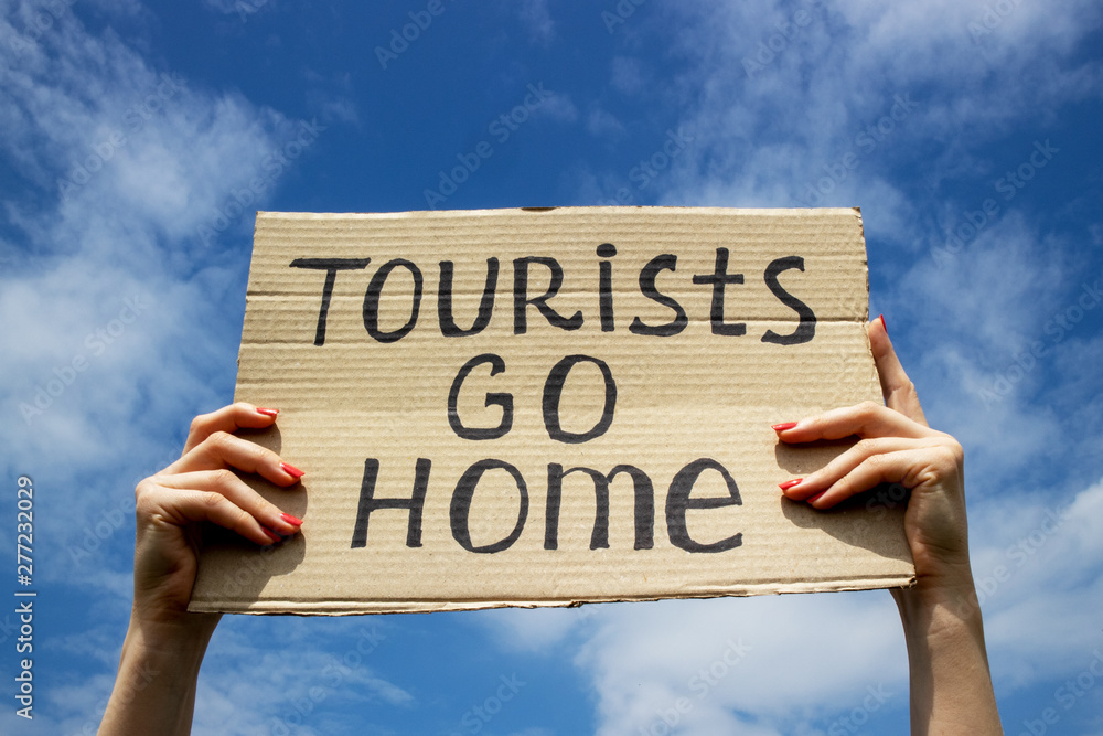 Text Tourists go home on a cardboard plate in hands. Blue sky with clouds background. Stop mass tourism concept. Text Tourists go home on a cardboard plate in hands. Blue sky with clouds background. Stop mass tourism concept.
