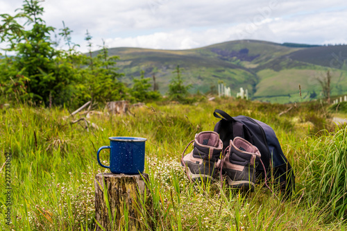 Selective focus on a camping blue enamel mug with hot coffee on a tree stump next to a pair of hiking boots and a backpack in mountain background. Adventure, travel and outdoor summer tourism concept.