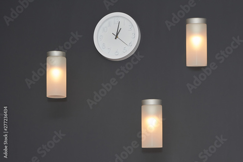 The white clock and three sconce lamps on gray wall. Style interior background.