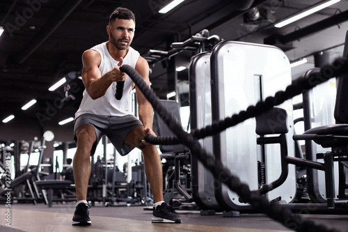 Fotografie Athletic young man with battle rope doing exercise in functional training fitness gym
