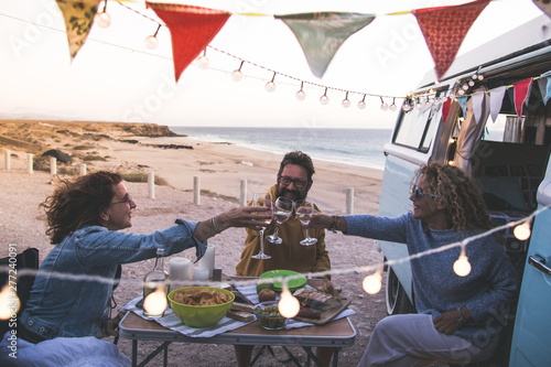 Happy friends cheering and drinking wine during camping vacation at the beach with vintage van. People having fun at weekend summer evening with camper. Youth lifestyle togetherness and party concept