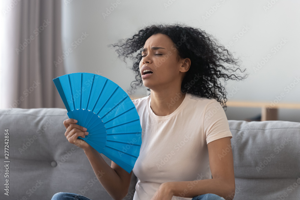 Overheated african young woman feeling hot waving fan at home - obrazy, fototapety, plakaty 