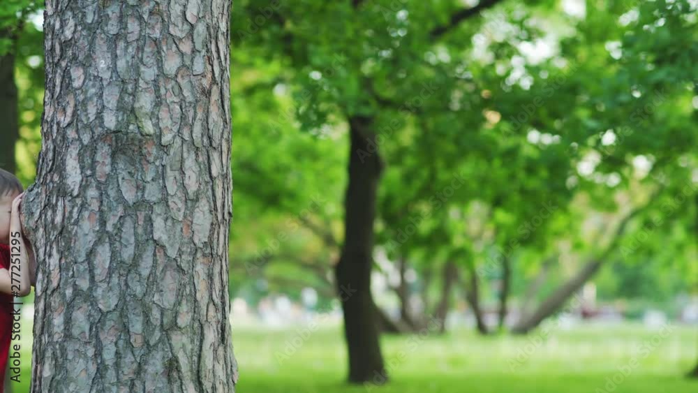 Group of kids playing hide and seek in park on summer day. Boy standing by tree trunk with closed eyes and counting, girls running to hide, tracking shot Group of kids playing hide and seek in park on summer day. Boy standing by tree trunk with closed eyes and counting, girls running to hide, tracking shot