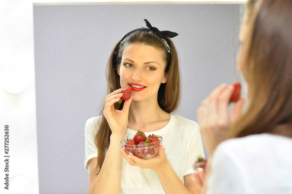 Beautiful sexy pin-up woman with bright makeup and red lipstick is eating red strawberries infront of the mirror and smiling. Beautiful sexy pin-up woman with bright makeup and red lipstick is eating red strawberries infront of the mirror and smiling.