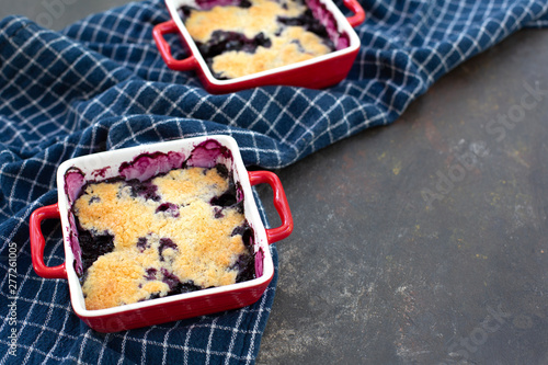 Fotografija Two freshly baked blueberry cobblers in red pans on blue kitchen towel with black textured background