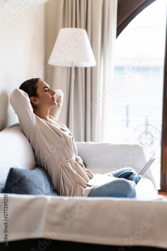 Pretty relaxed young woman sitting on white couch with hands over head with laptop computer photo