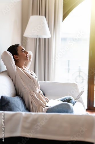 Pretty relaxed young woman sitting on white couch with hands over head with laptop computer photo