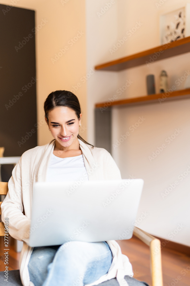 Young smiling brunette girl is sitting on modern chair in light cozy room at home working on laptop in relaxing atmosphere Young smiling brunette girl is sitting on modern chair in light cozy room at home working on laptop in relaxing atmosphere