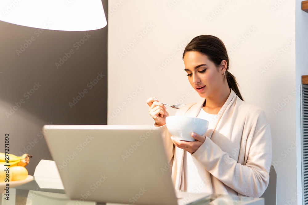Young woman having healthy breakfast in kitchen and looking at her laptop. Young woman in kitchen eating breakfast with laptop on counter. Young woman having healthy breakfast in kitchen and looking at her laptop. Young woman in kitchen eating breakfast with laptop on counter.