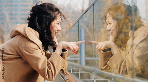 Beautiful Chinese girl with her mirror image in glass. Outdoor fashion portrait of glamour young Chinese cheerful stylish lady, emotions, people, beauty and lifestyle concept.
