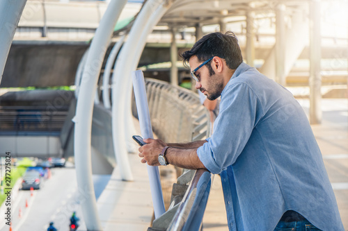 businessman standing at outside coridor using smartphone with one hand hold engineer plan photo