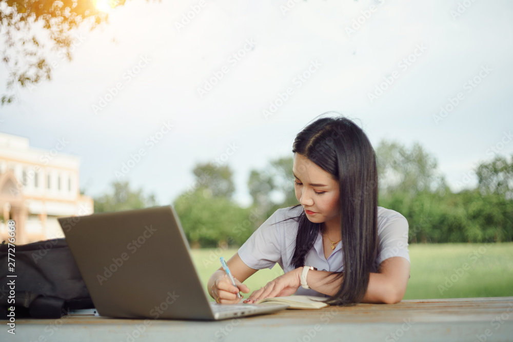 A young female university student writes a note and assignment with a laptop beside outside the campus with blure green tree background. Students self learning outside the campus. A young female university student writes a note and assignment with a laptop beside outside the campus with blure green tree background. Students self learning outside the campus.