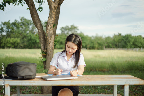 A young female university student writes a note and assignment with a laptop beside outside the campus with blure green tree background. Students self learning outside the campus.