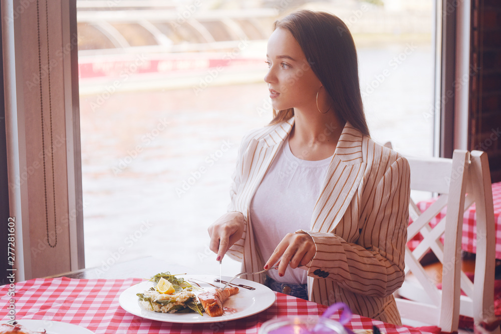 Brunette girl with long hair in a restaurant on ship on the background of the river. Girl tries salmon with rice. Girl eats appliances. The girl in the jacket after work eats. Brunette girl with long hair in a restaurant on ship on the background of the river. Girl tries salmon with rice. Girl eats appliances. The girl in the jacket after work eats.
