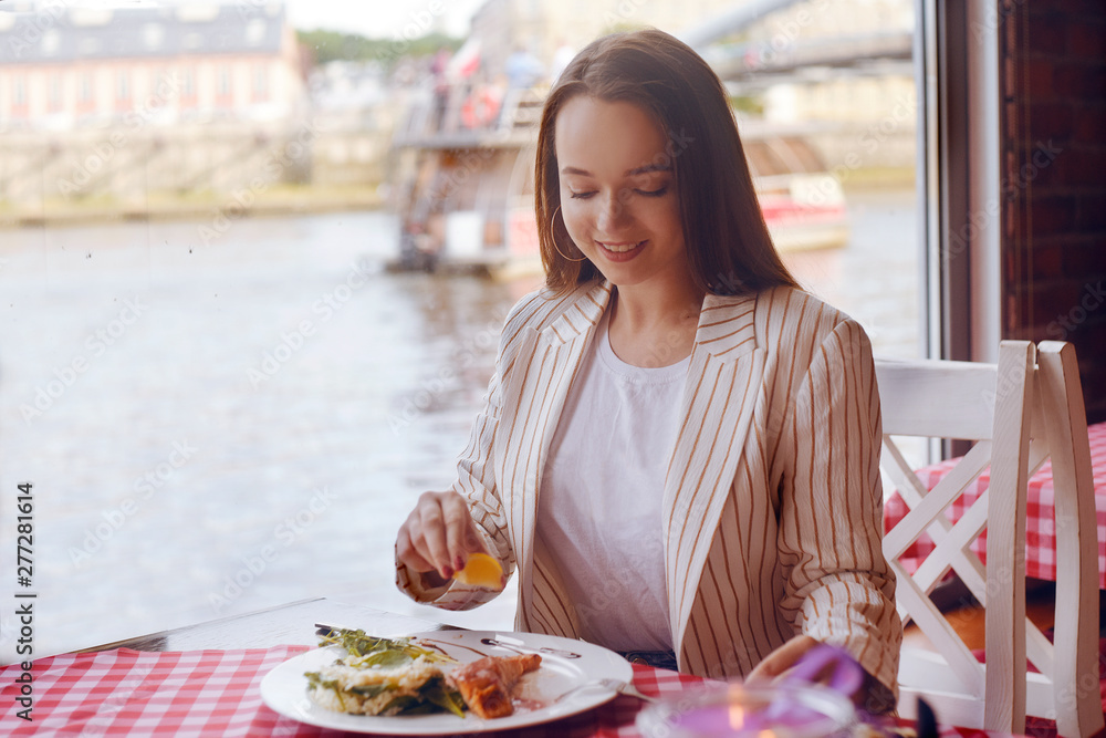 Brunette girl with long hair in a restaurant on ship on the background of the river. Girl tries salmon with rice. Girl eats appliances. The girl in the jacket after work eats.