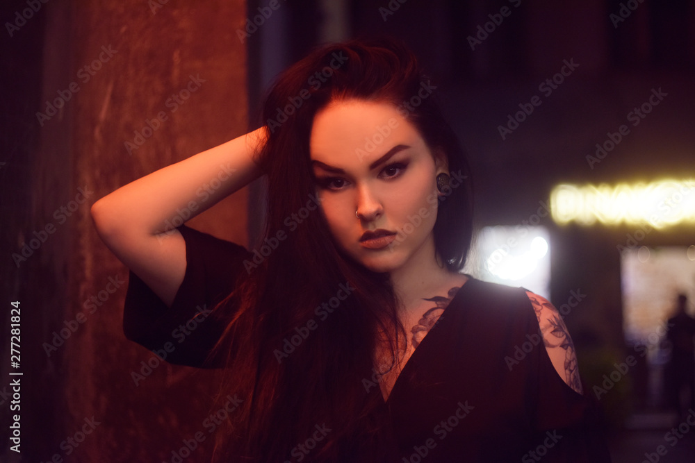Fototapeta premium The girl in the tattoo, with piercing in the nose , tunnels in the ears. Beautiful girl, night portrait. The girl in neon lighting. Beautiful unconventional brunette. Girl with a professional make-up.