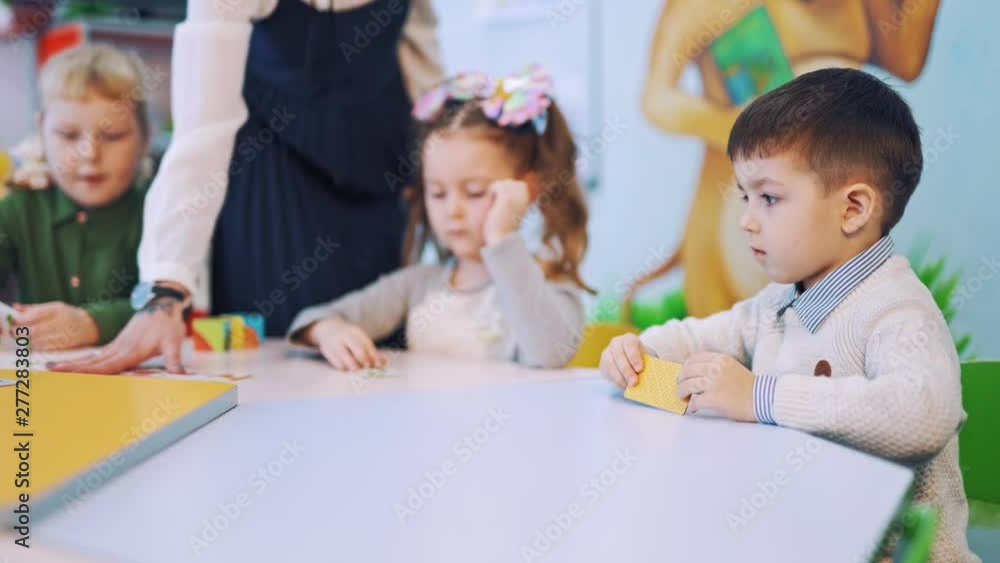 Little pupils are studying at elementary school. Teacher helps children to put picture cards into the necessary place on the table during the lesson. Little pupils are studying at elementary school. Teacher helps children to put picture cards into the necessary place on the table during the lesson.