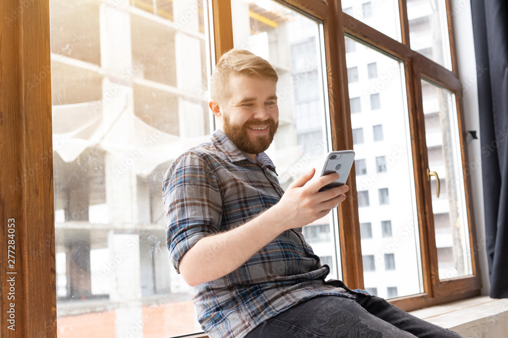 Fototapeta premium Cute young positive guy hipster with a beard is sitting on the windowsill by the big window and talking on the video communication using a smartphone and wireless high-speed Internet.