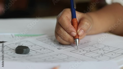 Hand of senior woman trying to solve sudoku puzzle with pencil as hobby on wooden office desk. Player insert numbers into grid consisting of nine squares subdivided into further nine smaller squares. photo