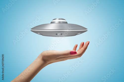 Side closeup of woman's hand and little UFO in air above her palm on light blue gradient background.