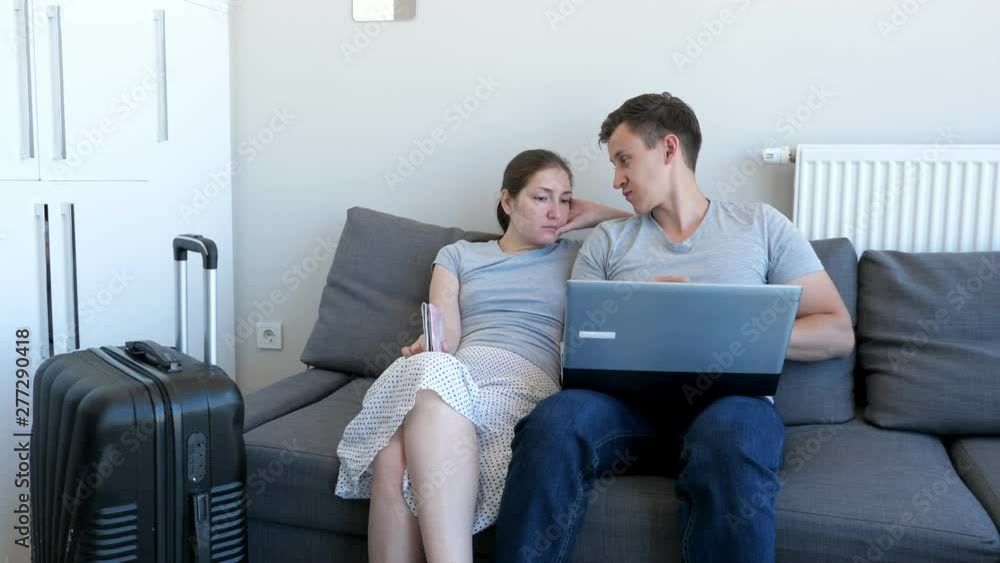 Young couple is booking transfer to the airport on laptop sitting on sofa at home. They are going on vacation with passports and suitcase. Choosing and booking tour and hotel. Young couple is booking transfer to the airport on laptop sitting on sofa at home. They are going on vacation with passports and suitcase. Choosing and booking tour and hotel.