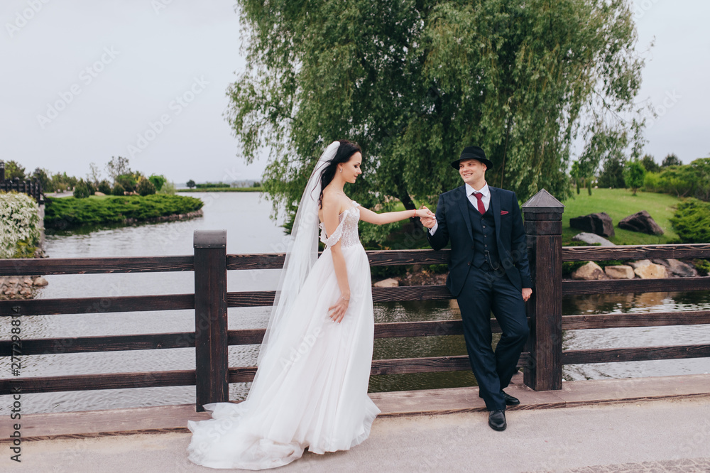 Beautiful newlyweds stand holding hands on an old bridge, against the backdrop of nature and the lake. Wedding portrait of a stylish groom in a hat and a beautiful brunette bride with a long dress.