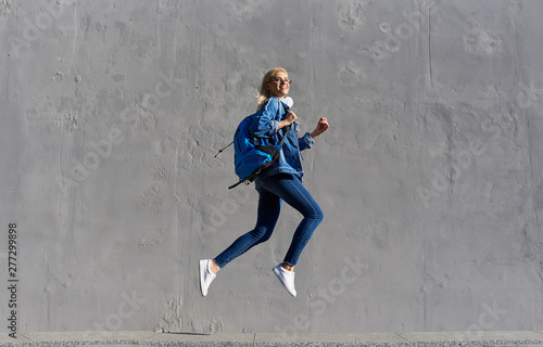 Full body Portrait of happy young woman walking with bag and cellphone. Cheerful student jumping in air over gray background