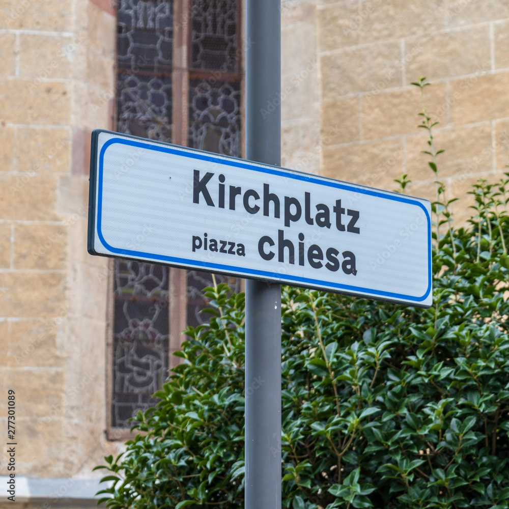 Bilingual Signs lettering Church Square, in german, Kirchplatz, and italian, piazzo chiesa. Church wall in background. Bilingual Signs lettering Church Square, in german, Kirchplatz, and italian, piazzo chiesa. Church wall in background.