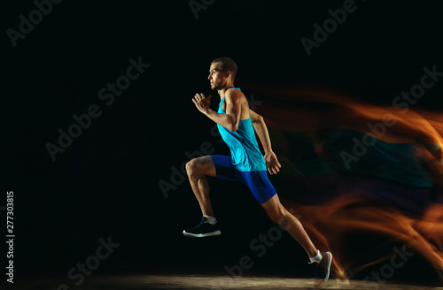 Professional male runner training isolated on black studio background in mixed light. Man in sportsuit practicing in run or jogging. Healthy lifestyle, sport, workout, motion and action concept.