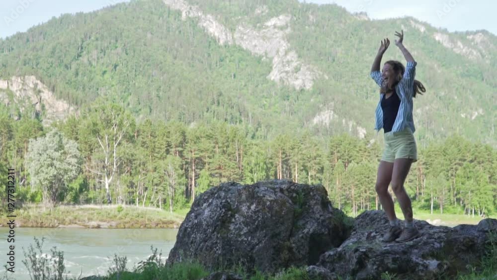 Slow shooting. The joy of climbing and overcoming the route. Happy woman jumping up and raising her arms standing on a rock by the mountain river Katun Slow shooting. The joy of climbing and overcoming the route. Happy woman jumping up and raising her arms standing on a rock by the mountain river Katun