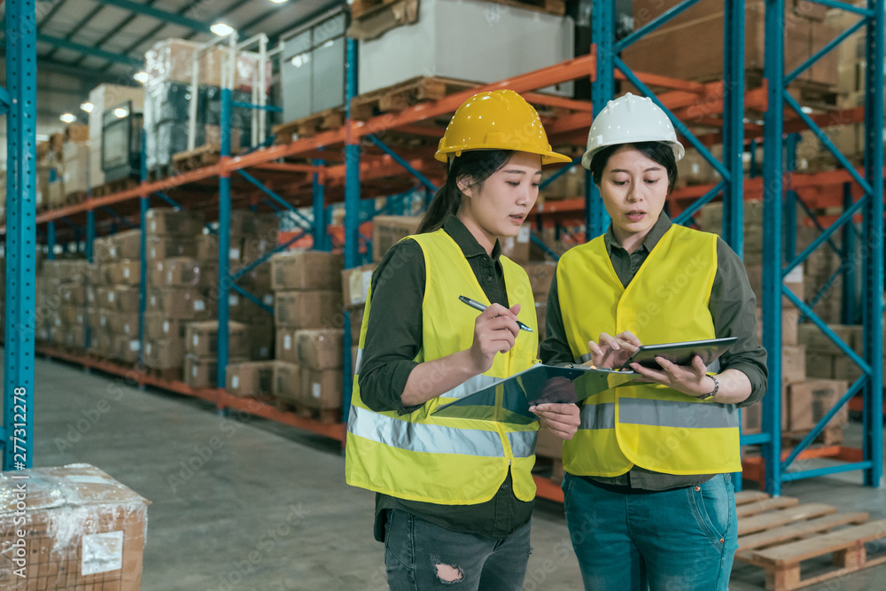 group of concentrated female workers using digital tablet in warehouse. two girl employees in helmet and safety vest discussing how to work in large stockroom. partners teamwork lifestyle concept. group of concentrated female workers using digital tablet in warehouse. two girl employees in helmet and safety vest discussing how to work in large stockroom. partners teamwork lifestyle concept.
