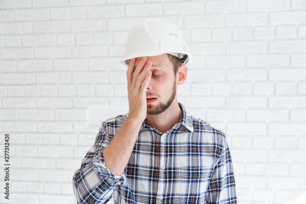 A blue collar worker wearing a hardhat is covering his face with his hand A blue collar worker wearing a hardhat is covering his face with his hand