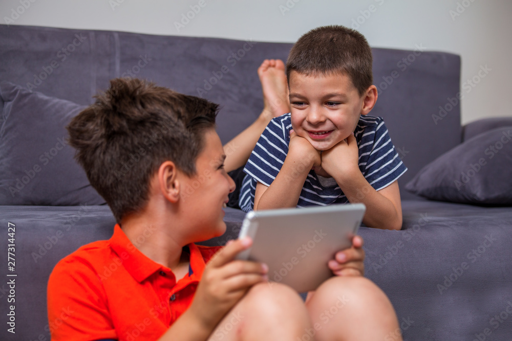 Pretty little boys are using a digital tablet, looking at each other and smiling while lying on the floor at home. brothers with tablet Pretty little boys are using a digital tablet, looking at each other and smiling while lying on the floor at home. brothers with tablet