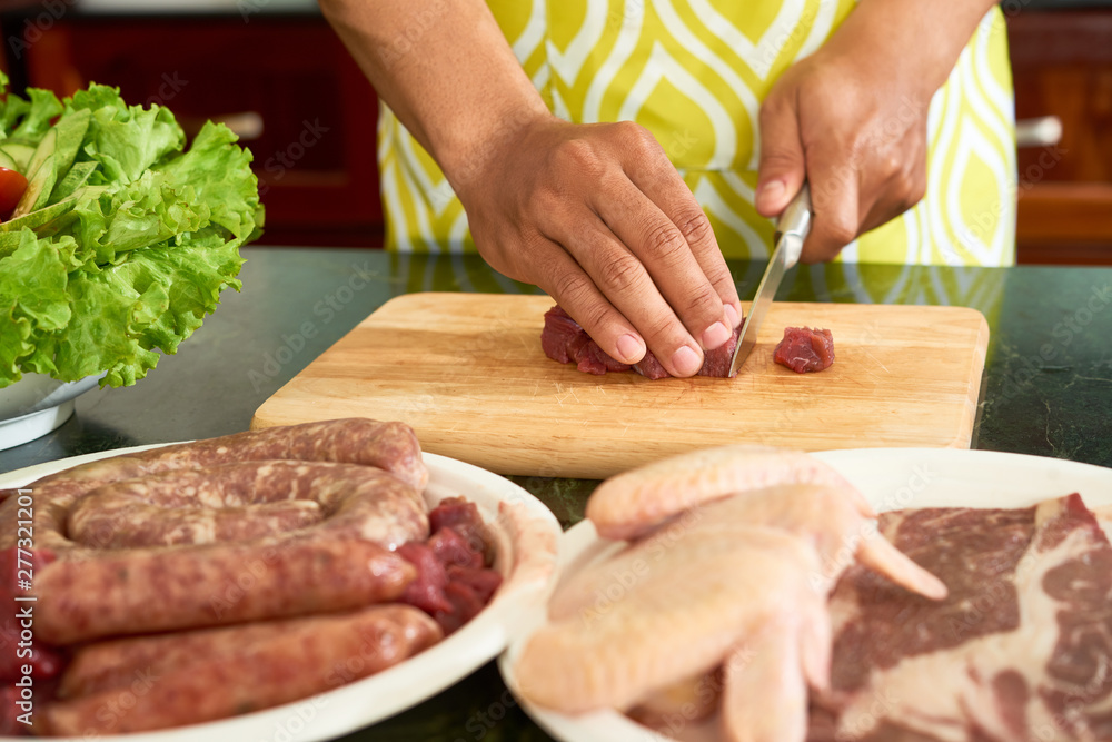 Close-up of young man cutting red raw meat on pieces with kitchen knife on cutting board in domestic kitchen