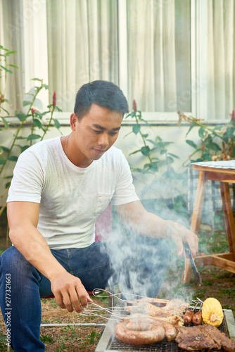 Asian young man sitting near the grill and cooking meat on it for lunch outdoors