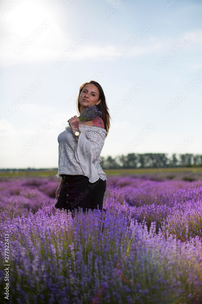 Young, beautiful woman with long hair and tattoos. Girl holding a fragrant bouquet and standing in a field among colorful, blooming lavender flowers in the summer in the countryside. Blue sky.