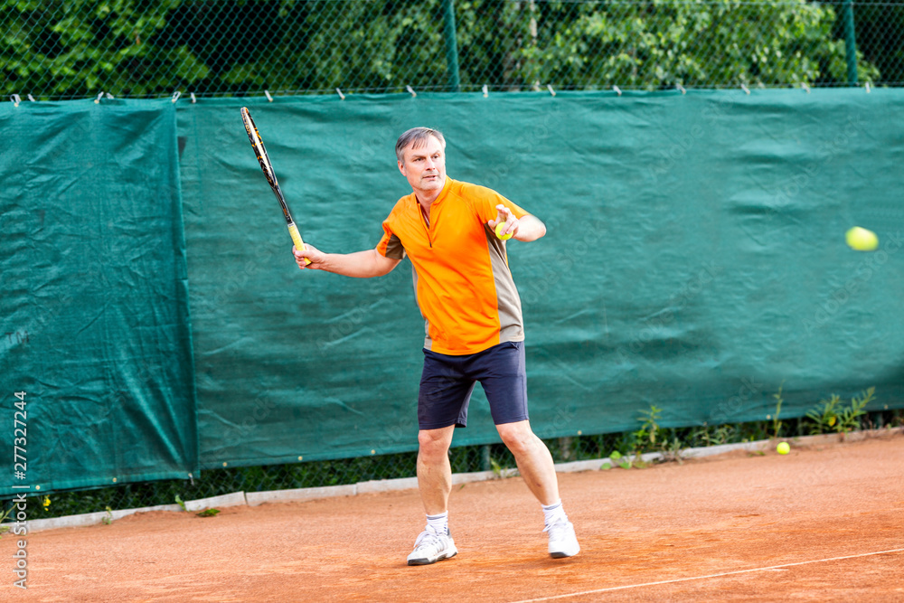 A middle-aged man plays tennis on a court with a natural earth surface on a sunny summer day.